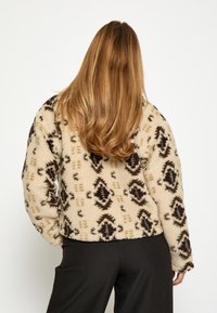 Bēžs fl�īsa žakets ar melniem un brūniem cilts rakstiem, kas piedāvā garas piedurknes un brīvu piegriezumu. Mīksta tekstūra, ikdienišķs dizains.