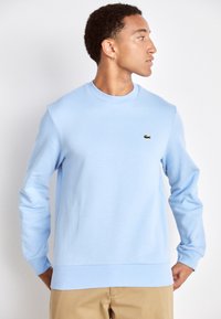 Sweatshirt bleu clair en tissu doux, avec un col rond, un ourlet côtelé et un petit logo vert Lacoste sur la poitrine.