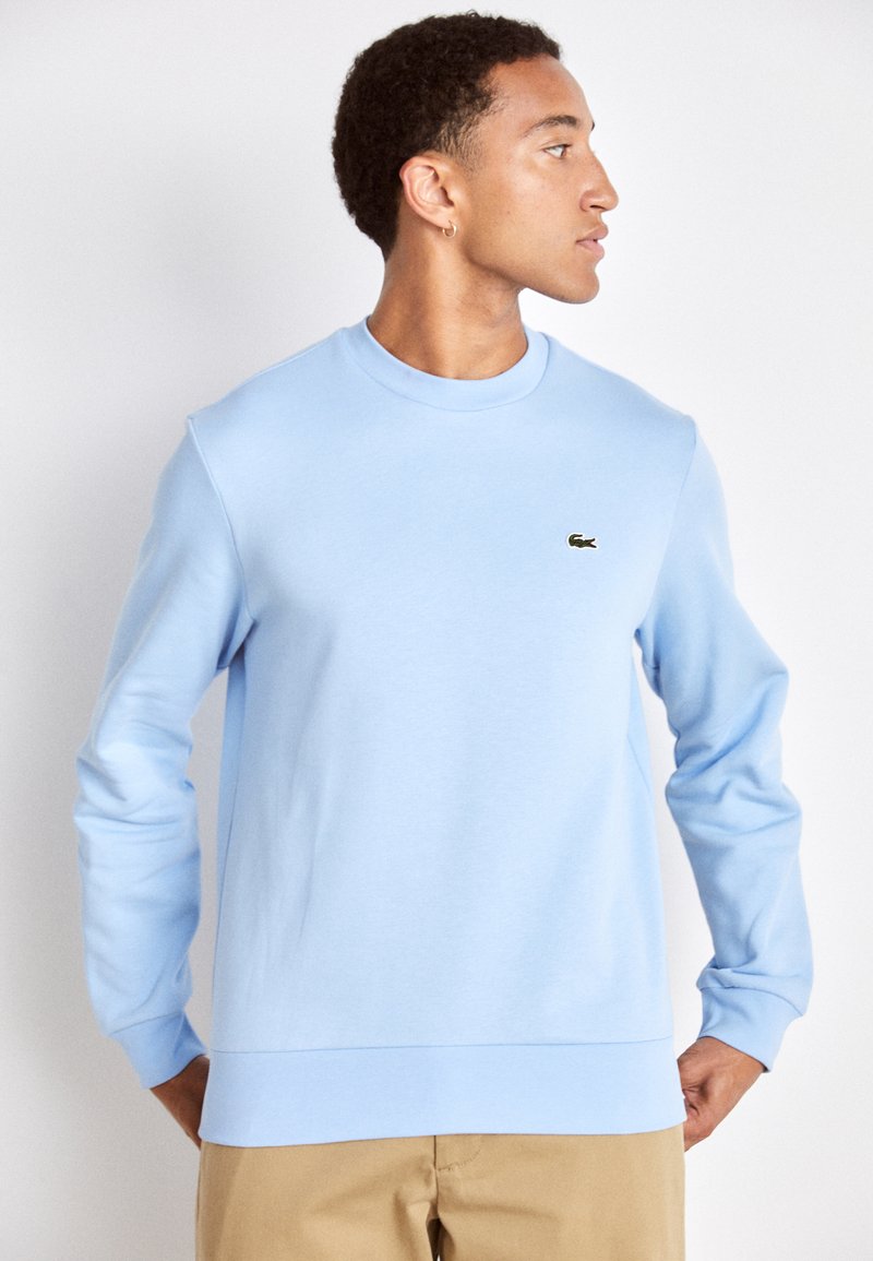 Sweatshirt bleu clair en tissu doux, avec un col rond, un ourlet côtelé et un petit logo vert Lacoste sur la poitrine.