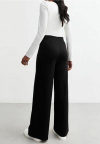 Pantalons large noirs en tissu doux, avec une taille haute, texture lisse, associés à un haut à manches longues blanc et des baskets.