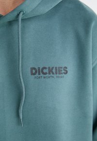 Sudadera con capucha de tela teal apagado que presenta una capucha, un bolsillo frontal y el logotipo negro "Dickies Fort Worth, Texas" impreso en el pecho.