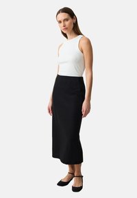 Jupe noire, à mi-mollet, ajustée, fabriquée en tissu lisse. Associée à un haut sans manches blanc et des chaussures plates noires. Design simple, sans motifs.