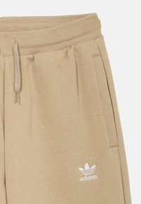 Béžové tepláky z mäkkej tkaniny s elastickým pásom. Majú predné vrecká a biele vyšívané logo Adidas na ľavej strane.