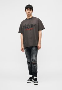 Γκρίζο γραφικό t-shirt με μαύρο και κόκκινο κείμενο, συνδυασμένο με μαύρο τζιν με σκισίματα και μαύρα και λευκά αθλητικά παπούτσια.