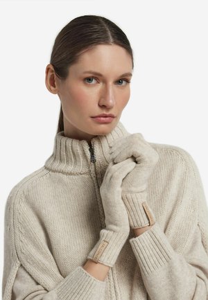 Beige gestrickte Handschuhe mit gerippten Bündchen, die eine strukturierte Oberfläche und ein Stoffetikett am Handgelenk aufweisen. Kombination mit einem Rollkragenpullover.