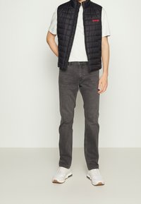 Czarna pikowana kamizelka bez rękawów na jasnoszarej koszulce, w zestawie z ciemnoszarymi dżinsami i białymi trampkami z niebieskimi i brązowymi akcentami.