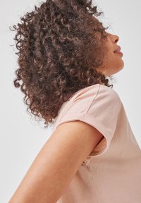 Žena s kudrnatými tmavými vlasy, oblečená ve světle růžovém tričku s krátkým rukávem, stojící bokem proti bezvýraznému bílému pozadí.