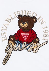 Eingesticktes Bärenmotiv auf einem weißen Stoff, der einen roten Pullover mit einem dreieckigen Logo trägt, umgeben von "ESTABLISHED IN 1981" und "guess" in Gold.