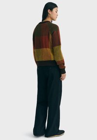 Maglione lavorato a maglia con audaci motivi a scacchi in rosso, marrone, giallo e verde. Collo e polsini a coste. Abbinnato a pantaloni larghi neri e scarpe.