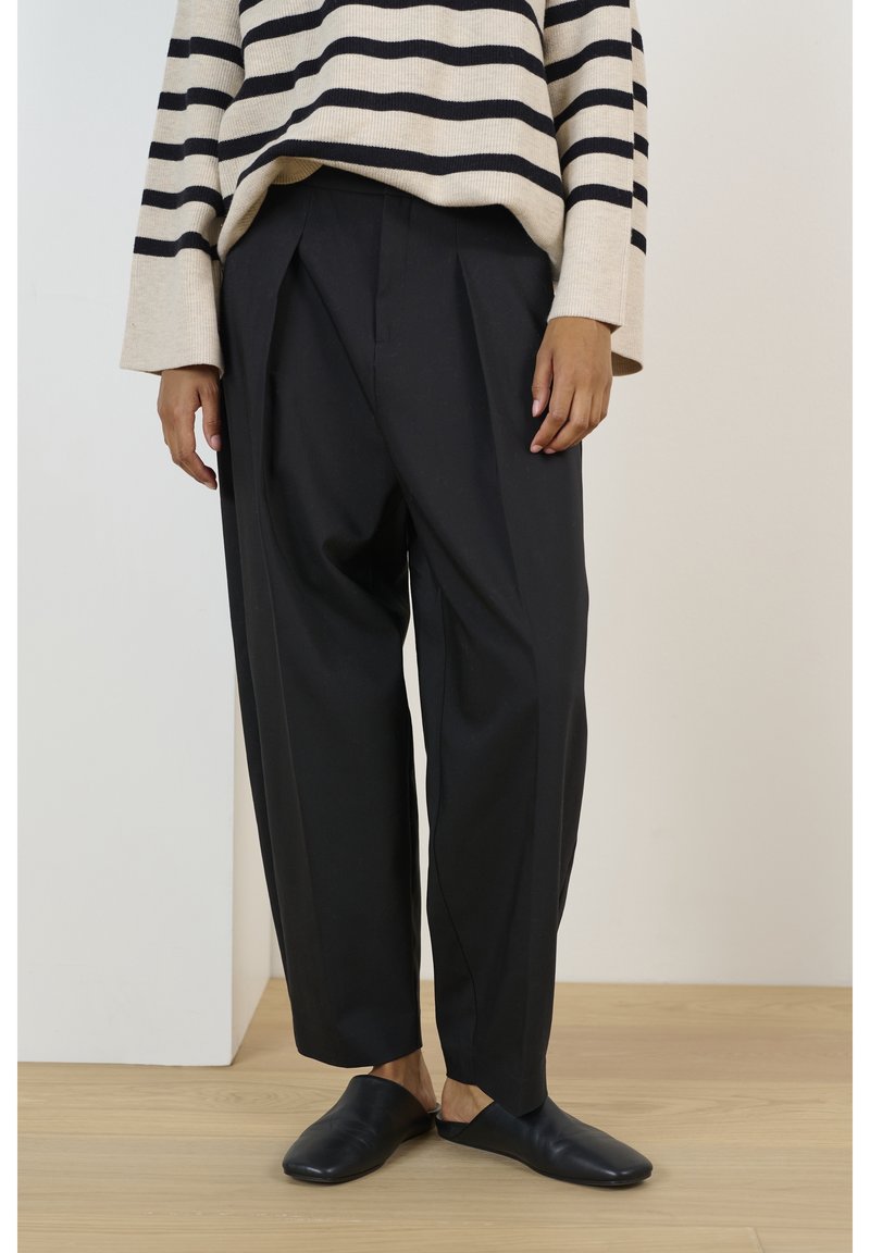 Personne portant un pantalon noir à jambes larges, des chaussures noires sans lacets et un pull beige à rayures horizontales noires, debout sur un sol en bois.