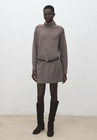 Pletené svetr šaty ve světle hnědé barvě s vysokým límcem a dlouhými rukávy, doplněné hnědými kozačkami po kolena a černým pásem.