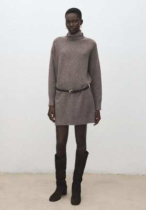 Vestido de punto en marrón claro, con un cuello alto y mangas largas, combinado con botas marrones hasta la rodilla y un cinturón negro.