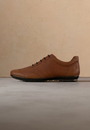 Baskets décontractées en cuir marron avec lacets et semelle noirs, vue latérale sur une surface grise devant un mur texturé beige.