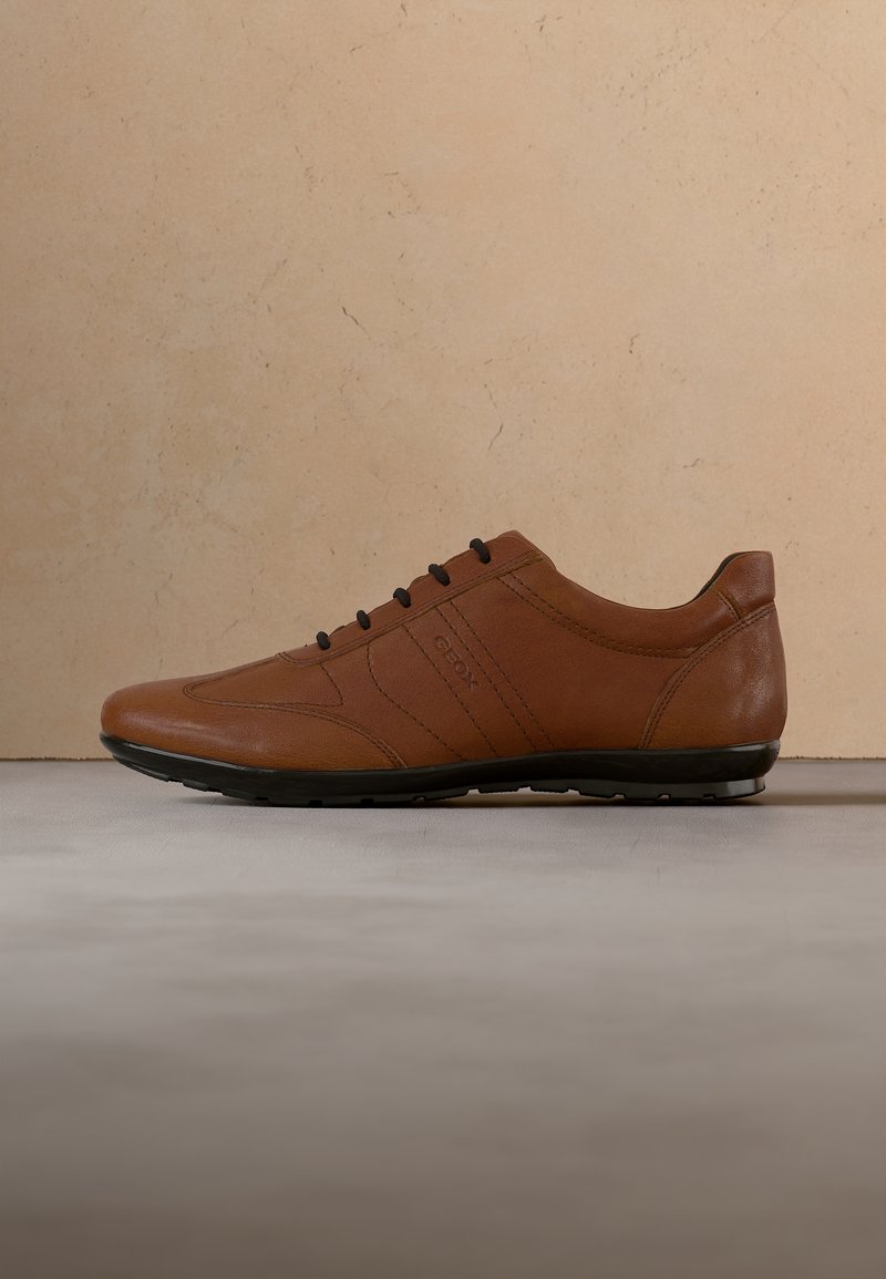 Baskets décontractées en cuir marron avec lacets et semelle noirs, vue latérale sur une surface grise devant un mur texturé beige.