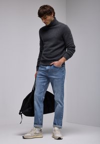 Suéter de cuello alto gris, jeans azul claro y zapatillas deportivas beige. Bolsa de deporte negra sostenida con una mano. Diseño simple y moderno.