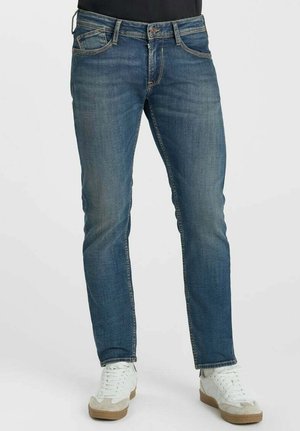 Mann trägt Slim-Fit blaue Jeans und weiße Sneakers, steht vor einem schlichten Hintergrund.