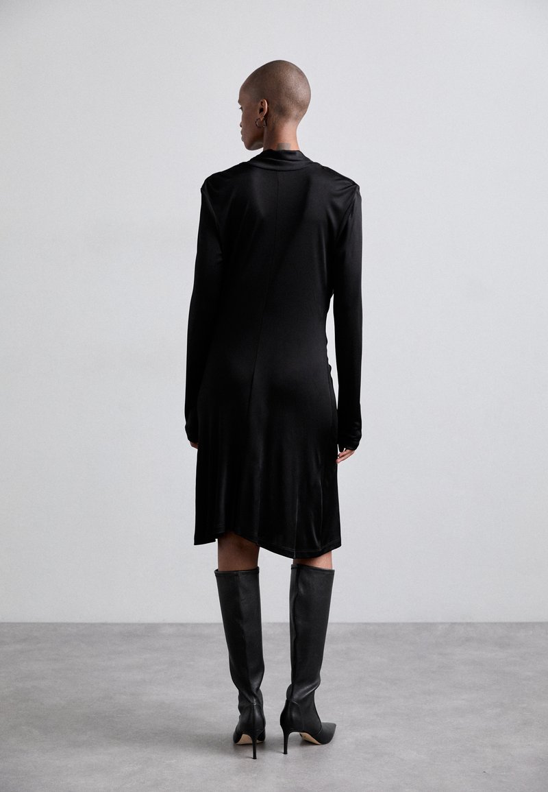 Black long-sleeve dress with a smooth texture, featuring a high neckline and an asymmetric hem. Paired with high, black knee-length stiletto boots.