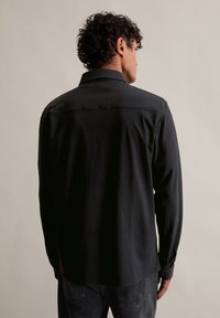 Čierna dlhá rukávová košeľa z bavlny, s golierom, gombíkovými manžetami a zakriveným lemom. Zadný pohľad zdôrazňuje jednoduchosť a čisté línie.