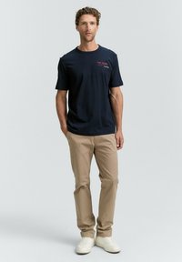 Navyblaue Baumwoll-T-Shirt mit kurzen Ärmeln, das ein rotes Textlogo aufweist. Kombiniert mit hellbeigen Hosen und weißen Turnschuhen. Einfaches, modernes Design.