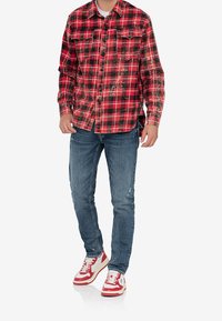 Rood en zwart geruite flanellen shirt met knoopsluiting aan de voorkant, twee borstzakken, versleten blauwe spijkerbroek en rood-witte sneakers.
