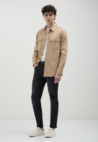 Beige Hemd mit Knöpfen und zwei Brusttaschen, kombiniert mit schwarzen, zerrissenen Jeans und weißen Sneakers. Glatte Textur und lässige Passform.