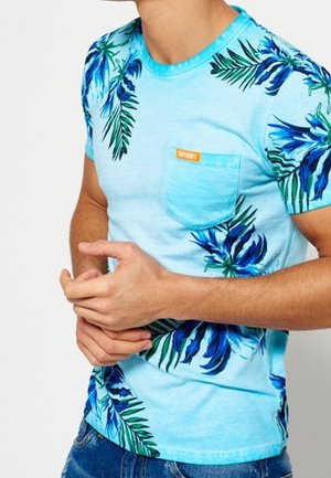 Mężczyzna w jasnoniebieskim tropikalnym t-shircie z zielonymi i niebieskimi liśćmi oraz małą przednią kieszenią, w połączeniu z niebieskimi dżinsami.