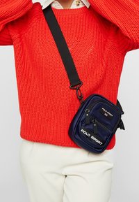 Borsa a spalla blu in nylon con cerniere, dotata di logo e tracolla regolabile, indossata sopra un maglione rosso lavorato a maglia.