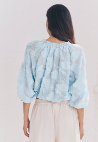 Blusa azzurro chiaro con texture e motivi floreali, scollo arricciato e maniche larghe, indossata sopra pantaloni color crema.