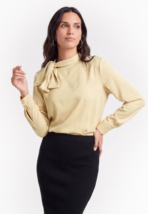 Frau mit langen dunklen Haaren, die eine beige Bluse mit Schleife und einen schwarzen, gerippten Rock trägt, posiert mit einer Hand auf der Hüfte und schaut nach links.