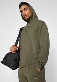 Olivengrønn hettegenser med snøring og svarte detaljer, kombinert med matchende joggebukser med gullbokstaver, holdt med en svart treningsbag.