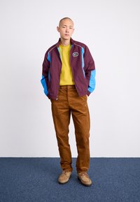 Mörk röd och blå vindjacka med en logotyp, gul skjorta, bruna kordbyxor och beige skor på en blåmatta.