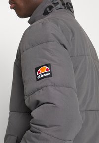 Grå pufferjakke med høj krave, der har et broderet logo-patch på ærmet og glat, struktureret stof.