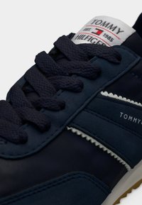 Σκούρο μπλε αθλητικό παπούτσι με ομαλή συνθετική επάνω επιφάνεια, κορδόνια από ύφασμα, λευκή επένδυση και ετικέτα στη γλώσσα με λογότυπο και κόκκινες και μαύρες λεπτομέρειες.