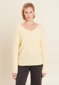 Helles gelbes V-Ausschnitt-Pullover aus weichem, strukturiertem Stoff mit überschnittenen Schultern und langen Ärmeln, kombiniert mit dunkelgrauen Hosen.