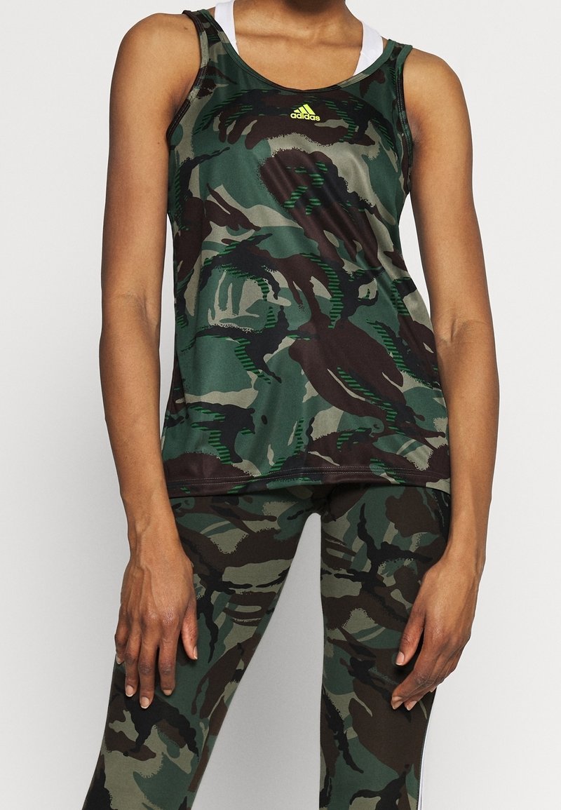 Personne portant un débardeur et un legging de camouflage vert et marron, debout devant un fond clair uni.