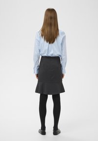 Chemise bleue à manches longues, jupe grise évasée, collants noirs et chaussures plates noires. Cheveux longs et raides, visibles de derrière.
