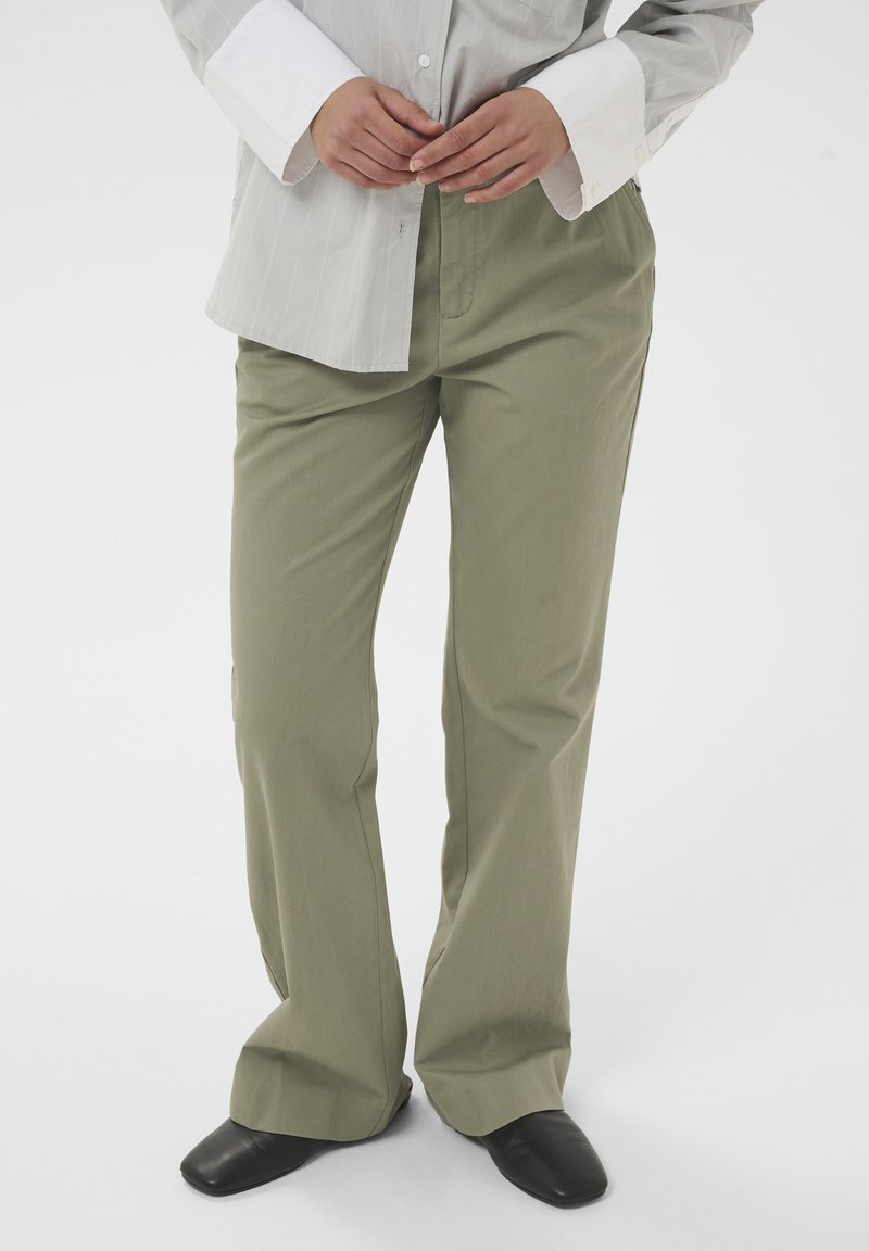 Personne portant un pantalon large de couleur olive, des chaussures noires à bout carré, et une chemise à manches longues gris clair avec des poignets blancs, debout devant un fond blanc.