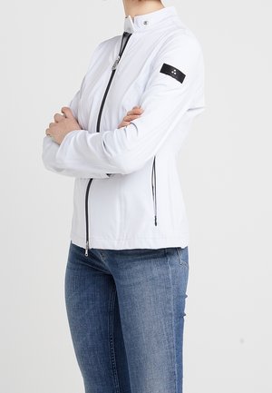 Person trägt weiße Jacke mit Reißverschluss und schwarzem Aufnäher am Ärmel sowie blaue Jeans, steht mit verschränkten Armen vor einem einfarbigen Hintergrund.