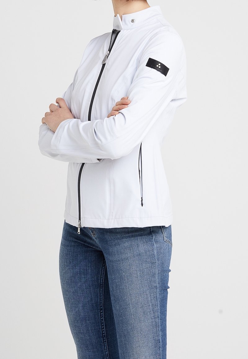 Personne portant une veste blanche zippée avec un patch noir sur la manche et un jean bleu, debout les bras croisés devant un fond uni.