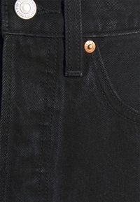 Gros plan de la poche avant d'un jean en denim noir avec un bouton argenté et des rivets en cuivre, montrant la texture du tissu et les coutures.
