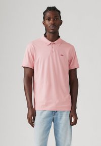 Polo rose en coton, avec un col à trois boutons et un petit logo rouge sur la poitrine. Associé à un jean bleu clair.
