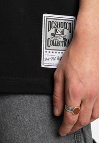 Černé tričko s bílou značkovou etiketou. Na etiketě je nápis "DC SHOE CO" a dekorativní prvky, doplněné šedými texturovanými kalhotami.