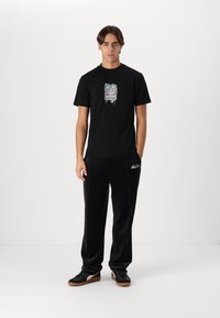Zwarte t-shirt met een grafisch ontwerp van een woeste masker, gecombineerd met zwarte velours broek en zwarte sneakers met bruine zolen.