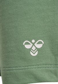 Grön bomullstyg med en strukturerad väv. En vit bi-logotyp är tryckt på den nedre delen, ovanför en sydd fåll.