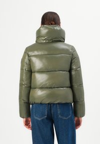 Persoon in een olijfgroene pufferjack met hoge kraag en blauwe jeans, van achteren weergegeven tegen een eenvoudige witte achtergrond.