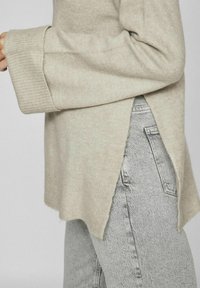 Pull en tricot beige avec des poignets côtelés et des fentes sur les côtés, associé à un jean déchiré gris clair. La texture semble douce et confortable.