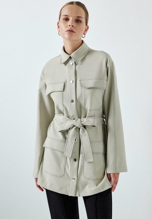 Femeie purtând o jachetă bej deschis cu centură, patru buzunare frontale și nasturi metalici argintii, asortată cu pantaloni negri, pe un fundal simplu.