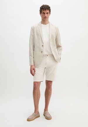 Mann, der einen beigefarbenen Blazer, ein weißes T-Shirt, beigefarbene Shorts und beigefarbene Slipper trägt, vor einem einfarbigen weißen Hintergrund.