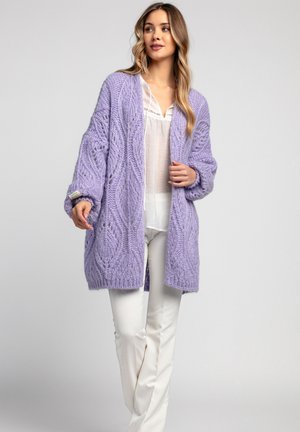 Fioletowy sweter dziergany w warkocze, z długimi rękawami i otwartym przodem. Nosi się go na białej bluzce, w połączeniu z białymi spodniami.