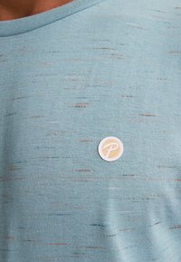 Light blue cotton T-shirt with a textured pattern, featuring small multicolored flecks and a round, white logo patch on the chest.